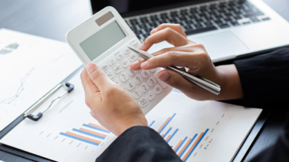 two hands using a calculator with a laptop in the background and a clipboard on the table