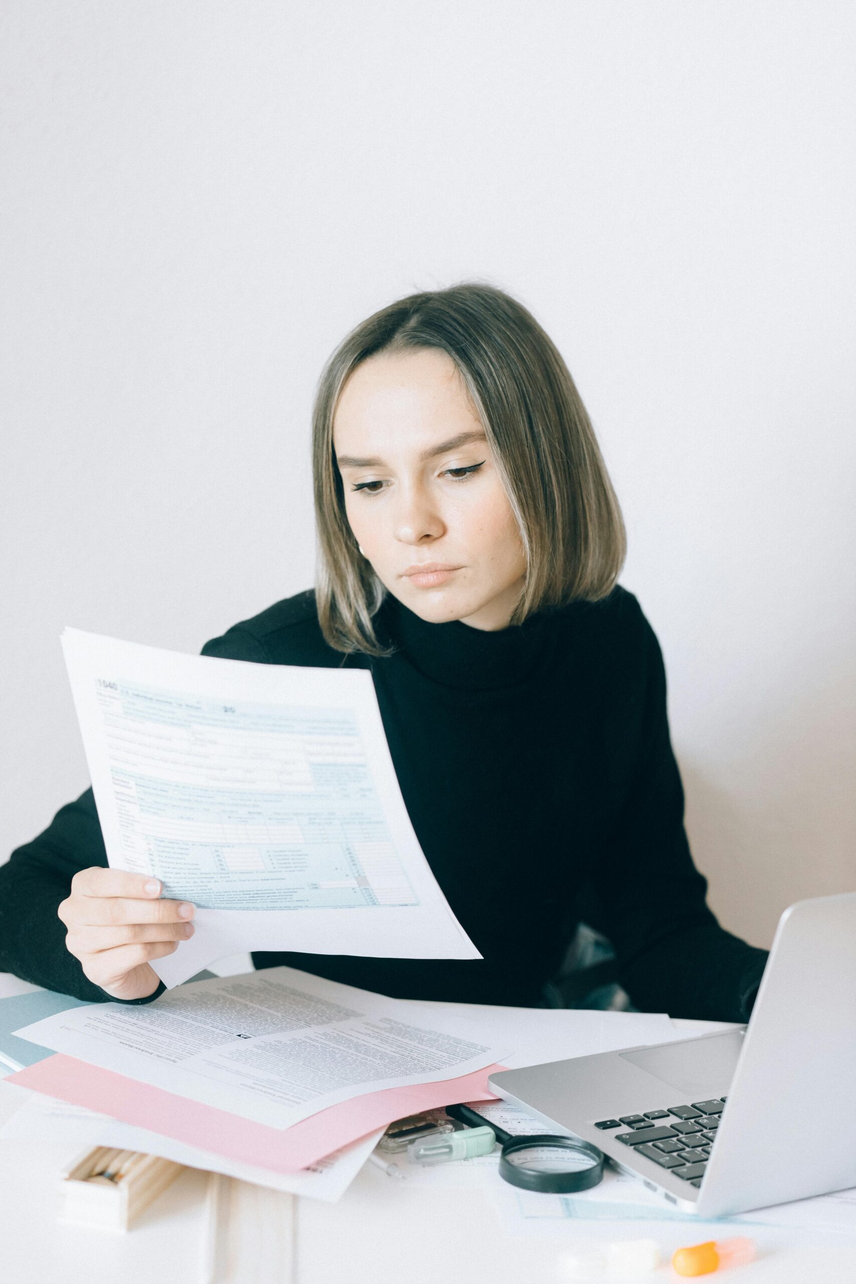 Focused woman reviewing paperwork at her desk with a laptop, showcasing a professional working environment.